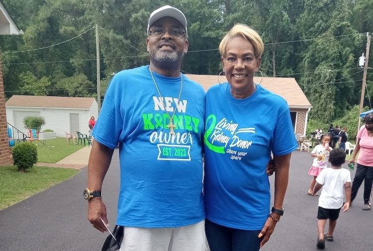 Two people standing in a neighborhood with blue shirts, one says that they received a transplant and the other says they’re the donor