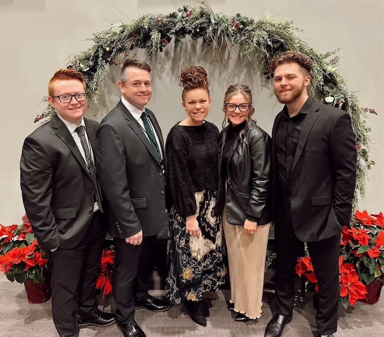 Family stand together in front of holiday wreath and flowers.